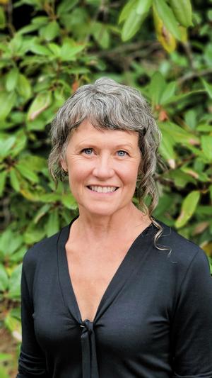  A headshot of Tracy smiling at you. She's wearing a dark grey blouse and standing in front of green foliage.